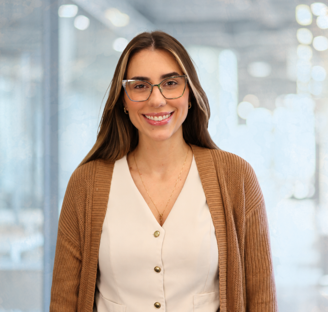 A woman wearing glasses and a tan cardigan over a white button-up top smiles at the camera.