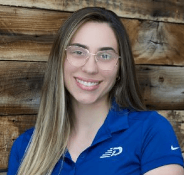 A woman wearing glasses and a blue polo smiles in front of a wooden wall.
