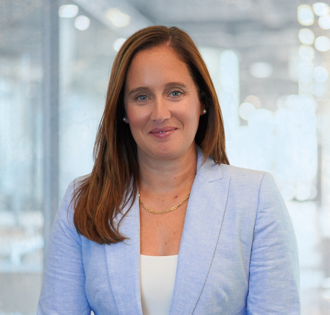 Caillin-Moye A woman with long brown hair wearing a light blue blazer and white top stands in a modern office setting, looking at the camera.