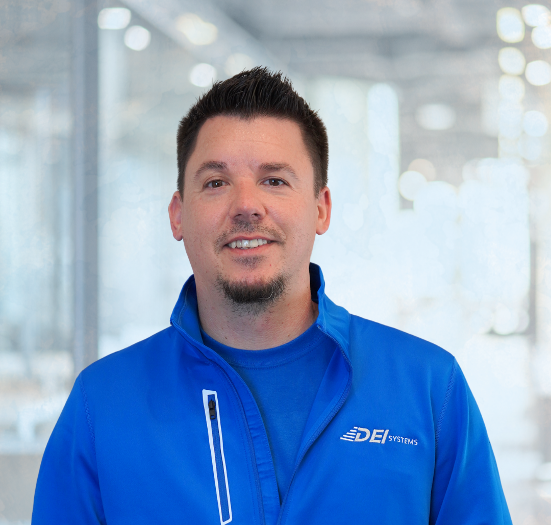 Kyle Hammond A man with short dark hair and a goatee, wearing a blue jacket and shirt with a “DEI Systems” logo, stands in a bright, modern office.