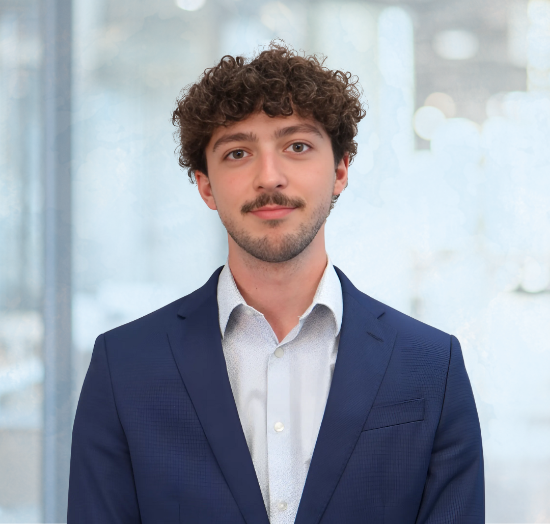 Matt Wentworth A young man with curly hair and a mustache wears a blue suit and white shirt, standing indoors in front of large windows with a blurred background.