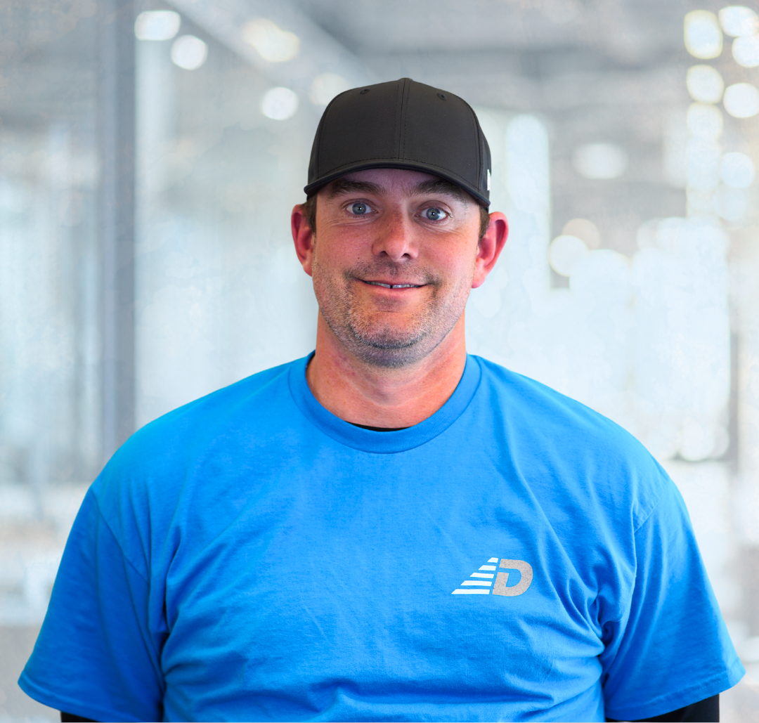 A man wearing a black cap and a blue t-shirt with a logo stands indoors in front of a blurred background.