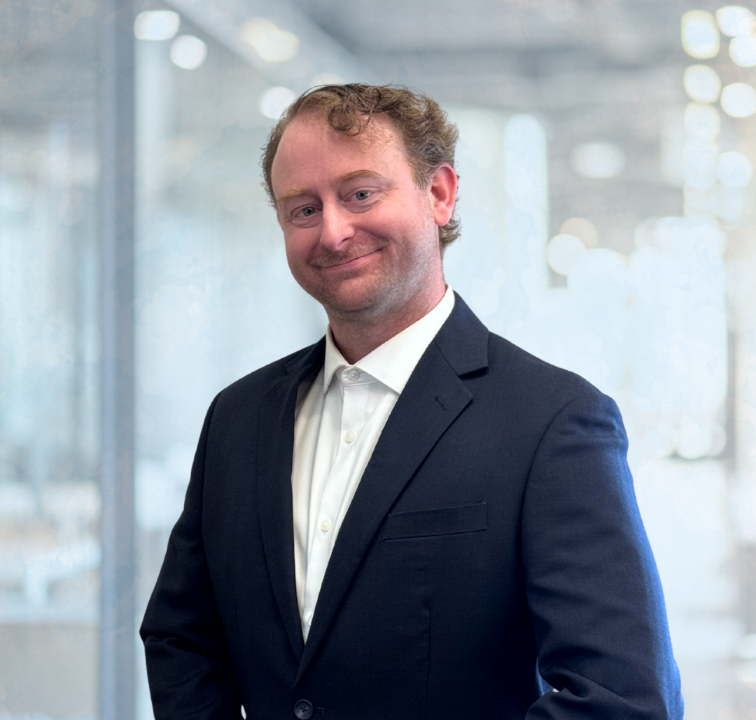 Corey Harwood Headshot A man in a dark suit and white shirt stands indoors with a slight smile, posing in front of a blurred office background.