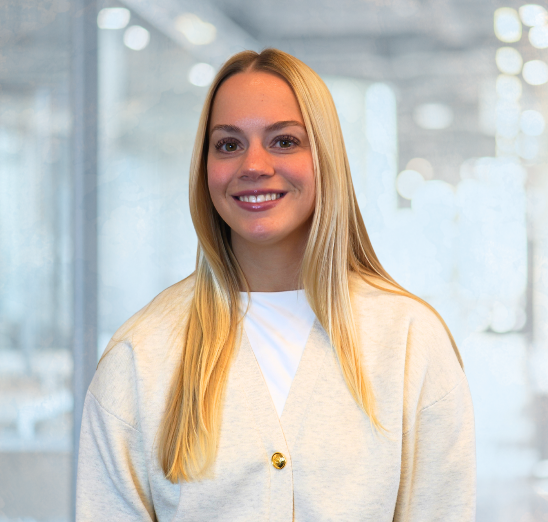 Alex_Final A woman with long blonde hair, wearing a light-colored buttoned cardigan and a white top, smiles in an office setting with glass walls and bright lighting.