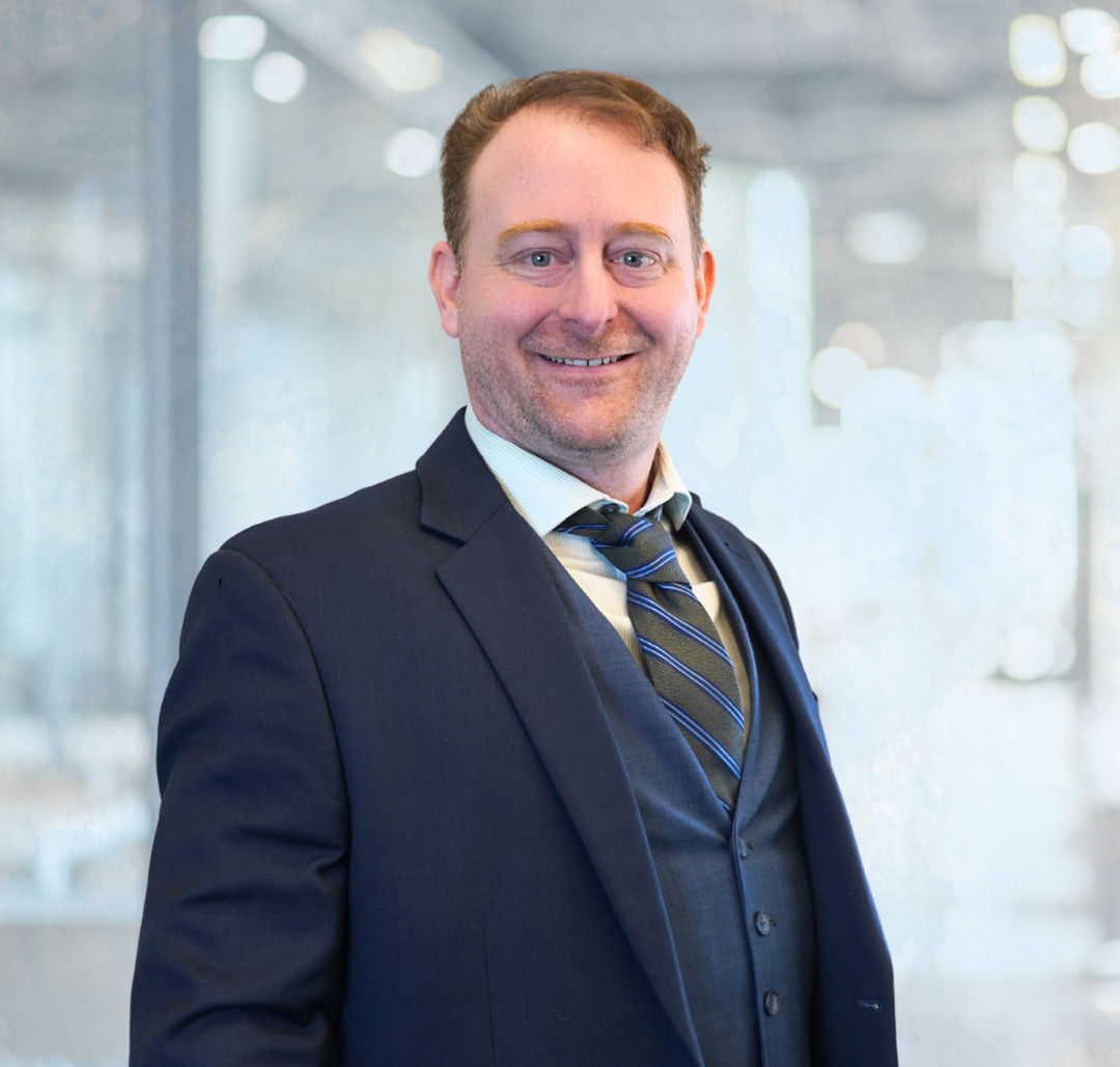 Corey_Final A man in a dark suit and tie stands indoors, smiling, with a blurred office background.