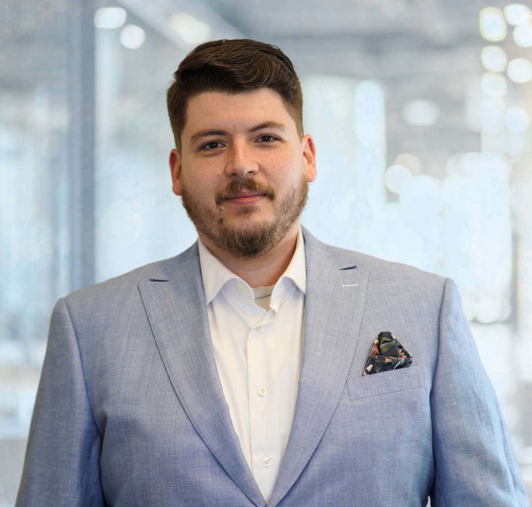 A man with short dark hair and a beard, wearing a light blue suit jacket and white shirt, stands indoors with a blurred office background.